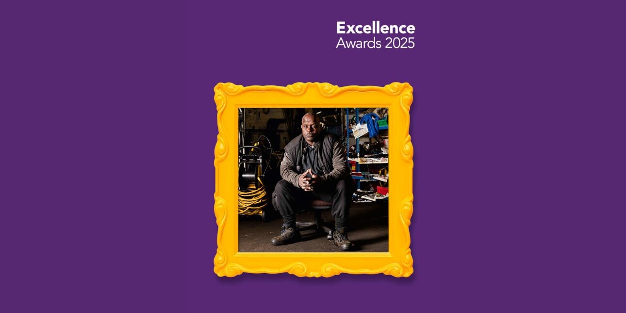 Superimposed on a purple background is a gold picture frame. Inside the frame is a photograph of a black man in black clothing sat in a workshop surrounded by tools and equipment.